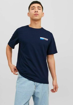 Marineblå bomulds T-shirt med korte ærmer, prydet med et brystlogo i hvid og blå. Modellen kombinerer det med lyseblå jeans.