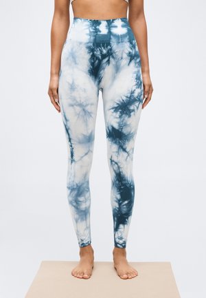 Vrouw die op blote voeten op een mat staat, gekleed in hoge taille legging met een blauwe en witte tie-dye print en naadloos ontwerp.