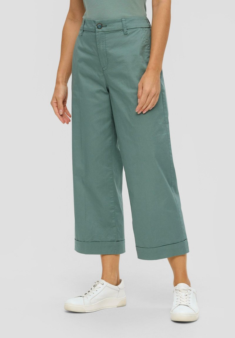 Groene wijde broek met een gladde textuur, voorzien van een knoopsluiting en omgeslagen manchetten, gecombineerd met witte sneakers.