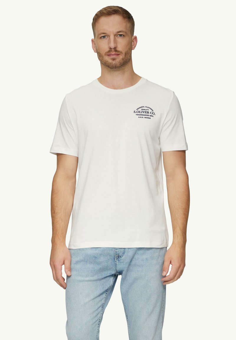 Weißes Baumwoll-T-Shirt mit kurzen Ärmeln, mit einem marineblauen Logo auf der linken Brust. Tragen mit hellblauen Jeans.
