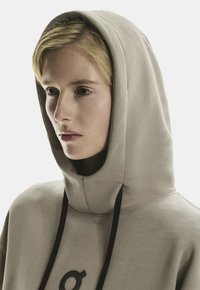 Hoodie beige con un ampio cappuccio, caratterizzato da lacci neri e un sottile dettaglio grafico. Texture liscia e vestibilità rilassata.