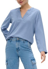Helle blaue Bluse mit V-Ausschnitt und langen Ärmeln, aus weichem Stoff. Hat eine lockere Passform und ein schlichtes Design. Wird mit hoch geschnittenen Jeans getragen.