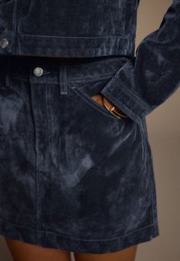 Ensemble veste et jupe en velours côtelé bleu foncé, avec tissu texturé, boutons en métal argenté et coutures apparentes sur les bords.