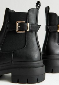 Botas negras hasta el tobillo con cuero suave y paneles laterales elásticos. Presentan una hebilla dorada en la correa y una suela texturizada.