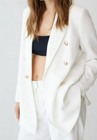 Witte dubbelzijdige blazer met gouden knopen, getailleerde pasvorm en textuurstof. Gedragen over een zwarte top met een gestructureerd ontwerp.