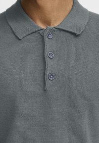 Grijze gebreide polo shirt met een zachte stof, een klassieke kraag en drie blauwe knopen op de sluiting. De textuur is soepel en gelijkmatig.