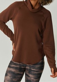 Bruine cowl-neck pullover met lange mouwen, gemaakt van zacht materiaal, gecombineerd met zwart en grijs gemusterde legging met een golvend ontwerp.