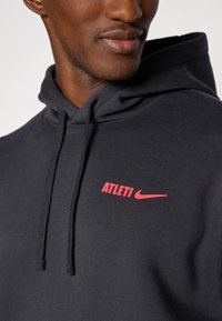 Crna dukserica s kapuljačom i vezicom, na kojoj se na lijevoj strani prsnog dijela nalazi crveni logotip "ATLETI" i Nikeov svastika. Mekana tekstura, ležeran dizajn.