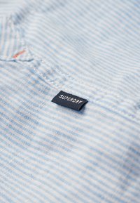 Lichtblauwe gestreepte stof met een gestructureerde afwerking. Bevat een marineblauwe label met "SUPERDRY" in witte letters. Close-up van de stikdetails.