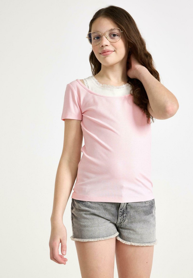 Lyserød ribbet t-shirt med korte ærmer og bred udskæring, lagdelt over en hvid top med blondekant. Parret med grå denim shorts.