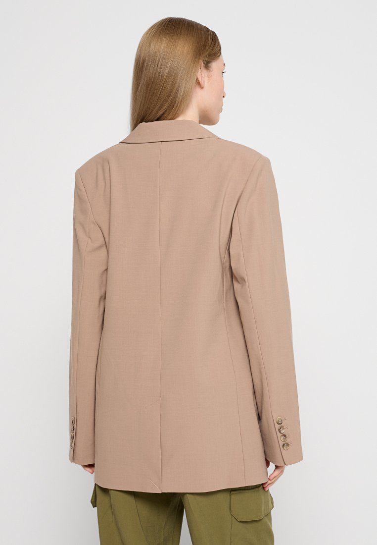 Donna con capelli biondi lisci, che indossa un blazer beige e pantaloni cargo verde oliva, vista da dietro su uno sfondo semplice.