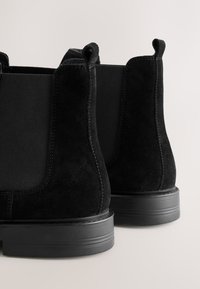 Melni zamšādas potīšu zābaki ar elastīgām sānu daļām un cilpām, ar plakanu, teksturētu gumijas zolīti un šuvju detaļām.