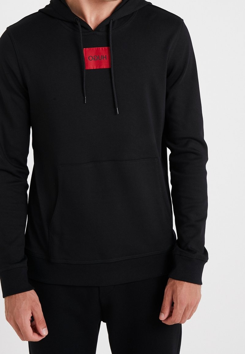Homme portant un sweat à capuche noir avec une poche chauve-souris à l'avant et une étiquette rectangulaire rouge comportant le texte "HUGO" en noir inversé.