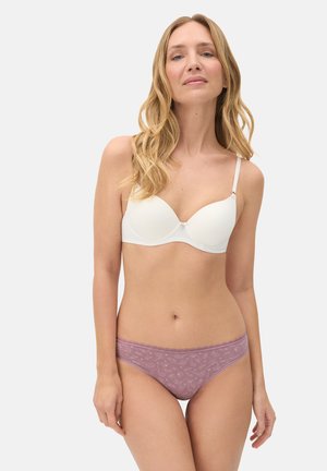 Mujer rubia que lleva un sujetador blanco ligeramente acolchado y bragas hipster lilas con estampado, de pie contra un fondo claro y liso.