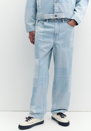 Muž nosí svetlomodré patchwork rifle a zodpovedajúcu bundu, s bielymi teniskami a čiernymi šnúrkami, stojí na bielom pozadí.