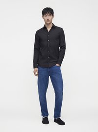 Jeune homme debout portant une chemise noire boutonnée, un jean bleu et des chaussures noires à enfiler sur un fond blanc uni.
