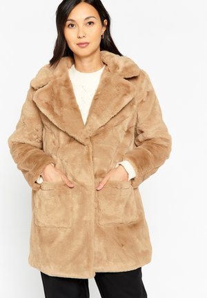 Manteau en fausse fourrure de couleur beige clair avec un grand col, des poches avant et une texture douce, porté sur un pull crème.
