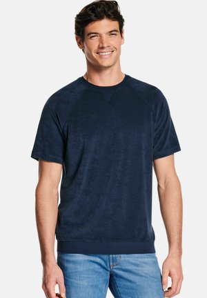 Homme souriant portant un sweat-shirt à manches courtes bleu marine et un jean bleu clair, sur un fond blanc uni.