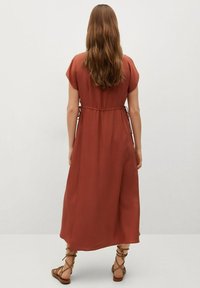 Frau mit langen, welligen braunen Haaren, die ein rostfarbenes Midi-Kleid mit seitlichen Bändern und braunen schnürlosen Sandalen trägt, steht vor einem schlichten Hintergrund.