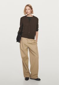 Brauner, geknöpfter Cardigan über einem schwarz-weiß gestreiften Hemd, kombiniert mit beigefarbenen, plissierten Hosen und schwarzen flachen Schuhen. Einfacher und lockerer Schnitt.