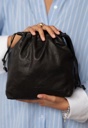 Person iført en blåstribet skjorte holder en sort skindtaske med snøre med begge hænder, hvor en guldring og et sølvarmbånd er synlige.