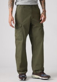 Cargo pants in olive green cotton, featuring side cargo pockets, straight cut, and contrasting stitching. Worn with dark athletic shoes.