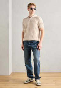 Light beige knit polo shirt with short sleeves, light structured texture. Blue jeans, green and cream-striped sneakers, and sunglasses.