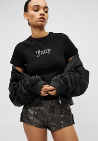 Μαύρο κοντό μπλουζάκι με τη λέξη "Juicy" σε ασημένιες στρας, συνδυασμένο με μαύρο μπουφάν τύπου puffer και μεταλλικά γκρι σορτς με διακοσμητικά κουμπιά.