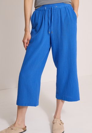 Personne portant un pantalon large bleu vif et court avec une taille à cordon, des chaussures beiges à enfiler et un haut gris, debout à l'intérieur.
