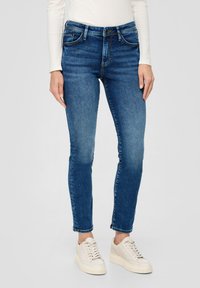 Cienkie dżinsy w średnim niebieskim kolorze, z wysokim stanem, pięcioma kieszeniami i delikatnym wyblaknięciem na udach. Noszone z jasnymi sneakerami.