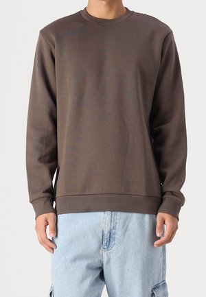 Person iført en ensfarvet brun langærmet sweatshirt og lyseblå jeans, vist fra skuldre til lår mod en ensfarvet baggrund.