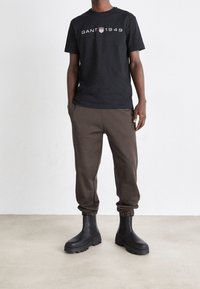 Camiseta negra con el logo "GANT 1949" en blanco, combinada con pantalones de chándal marrones y botas negras de tobillo. Ajuste casual con puños elásticos.