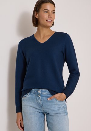 Femme portant un pull bleu marine à col en V et un jean bleu clair, debout avec une main dans la poche devant un fond uni.