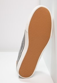Zapatilla de lona gris con suela de goma blanca y borde negro en contraste. La suela exterior es de un marrón texturizado con el nombre de la marca grabado.