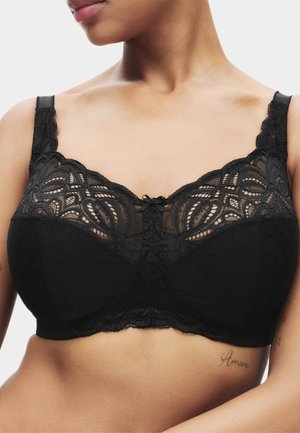 Femme portant un soutien-gorge noir en dentelle avec de larges bretelles en dentelle et des motifs floraux complexes sur les bonnets supérieurs.