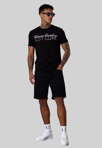 T-shirt noir avec texte blanc, associé à un short en denim noir avec des bords effilochés. Porté avec des baskets blanches et des lunettes de soleil noires.