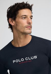 Marinblå T-shirt med långa ärmar i bomull, med broderad text "POLO CLUB" och "ESTABLISHED 2012" i vitt och rött på framsidan.