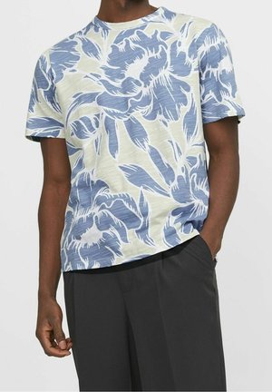 Mand iført hvid kortærmet t-shirt med stort blåt blomsterprint og sorte bukser, den ene hånd i lommen, mod ensfarvet baggrund.