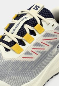 Zapatilla de correr gris con parte superior de malla texturizada, detalles en azul marino, lazos para los cordones amarillos, líneas decorativas rojas y media suela blanca.