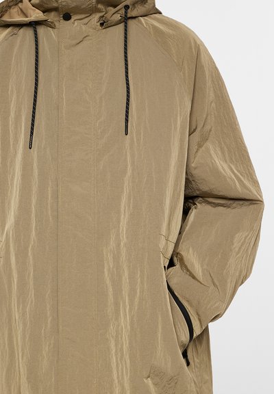 Beige huva jacka med en skrynklig textur, svarta dragsko och fickor med dragkedja. Den har en avslappnad passform och en minimalistisk design.