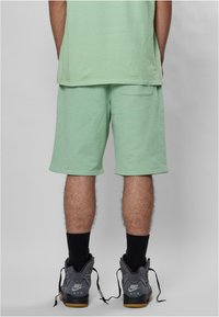 Shorts in cotone verde menta con vita elastica, tasca posteriore e vestibilità rilassata, abbinati a sneakers grigie Nike e calzini neri.