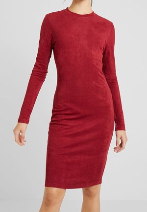 Frau trägt ein figurbetontes, langärmeliges, knielanges rotes Kleid aus strukturiertem Stoff und steht vor einem schlichten hellgrauen Hintergrund.