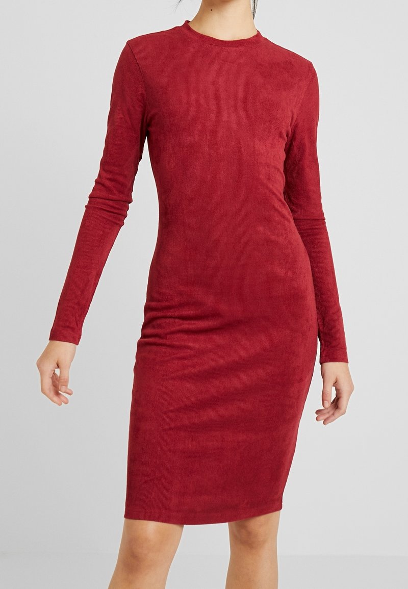 Femme portant une robe rouge moulante, à manches longues et longueur genou, en tissu texturé, debout devant un fond gris clair uni.
