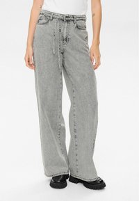 Personne portant un jean gris délavé taille haute et ample, avec une ceinture nouée et des chaussures noires imposantes, sur un fond blanc.