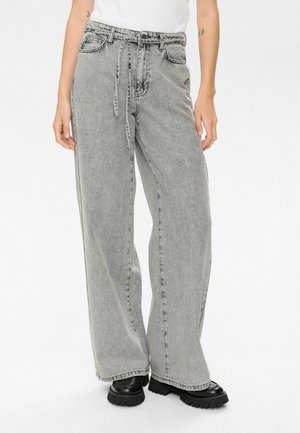 Person wearing high-waisted, loose-fitting grey washed jeans with a tied belt and black chunky shoes against a white background.