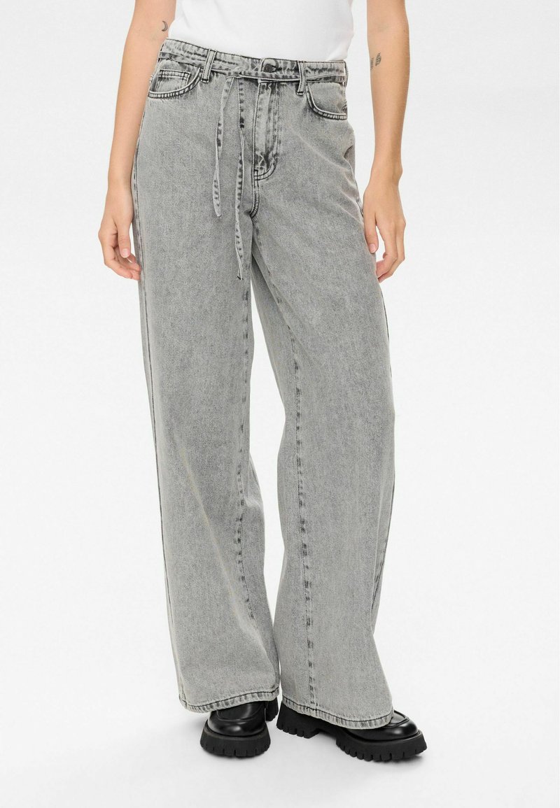 Personne portant un jean gris délavé taille haute et ample, avec une ceinture nouée et des chaussures noires imposantes, sur un fond blanc.