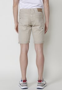 Person står vänd bort med beiga kortbyxor uppviktade, vita sneakers med röda och marinblå accenter, och en vit t-shirt mot en enfärgad bakgrund.