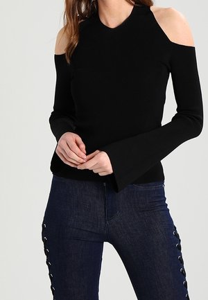 Kobieta ubrana w czarną bluzkę z odkrytymi ramionami i rozkloszowanymi rękawami oraz ciemnoniebieskie jeansy z bocznymi wiązaniami.