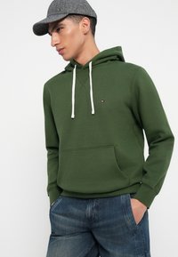 Zaļš kokvilnas hoodijs ar priekšējo kabatu, baltām auklām un mazu logotipu. Saskaņots ar plašiem ziliem džinsu šortiem. Pelēka cepure uz galvas.