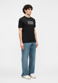 Čierne bavlnené tričko s bielym geometrickým vzorom na prednej strane, kombinované so svetlo modrými džínsami s širokými nohavicami a béžovými teniskami.